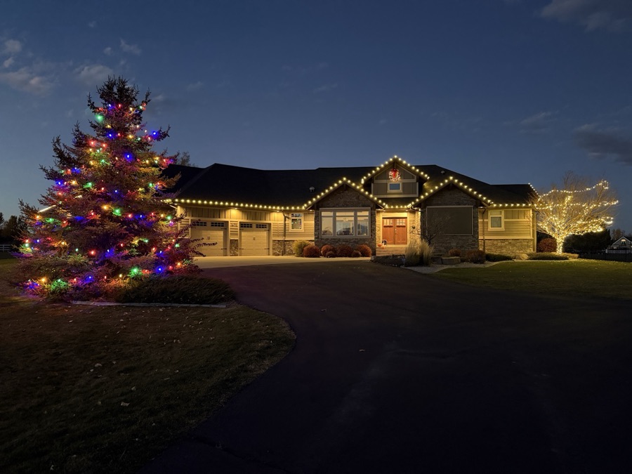 Ranch home with warm white roofline beside a large evergreen wrapped in multicolor lights at twilight